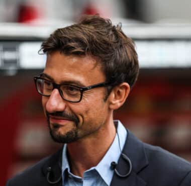 Man with dark hair and glasses, wearing a blue shirt and dark blazer, looking to the left outdoors.