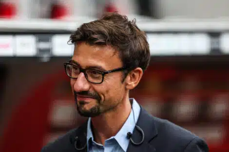 Man with dark hair and glasses, wearing a blue shirt and dark blazer, looking to the left outdoors.