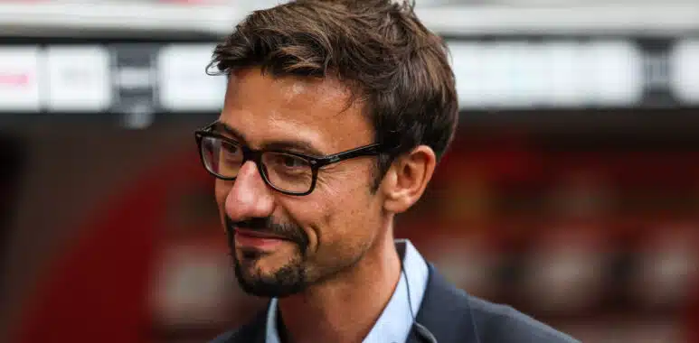 Man with dark hair and glasses, wearing a blue shirt and dark blazer, looking to the left outdoors.