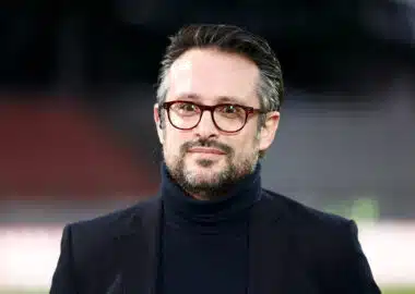 Man wearing a dark blazer and turtleneck with red-framed glasses, outdoors, smiling softly (close-up headshot)
