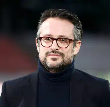 Man wearing a dark blazer and turtleneck with red-framed glasses, outdoors, smiling softly (close-up headshot)