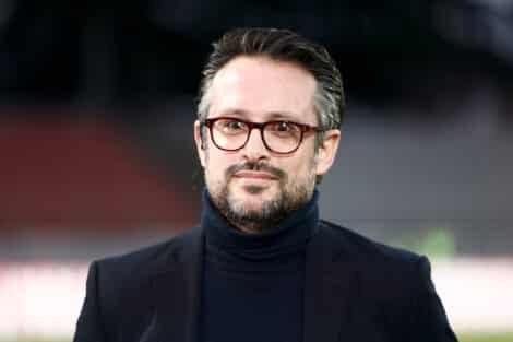 Man wearing a dark blazer and turtleneck with red-framed glasses, outdoors, smiling softly (close-up headshot)