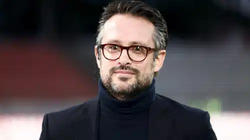 Man wearing a dark blazer and turtleneck with red-framed glasses, outdoors, smiling softly (close-up headshot)