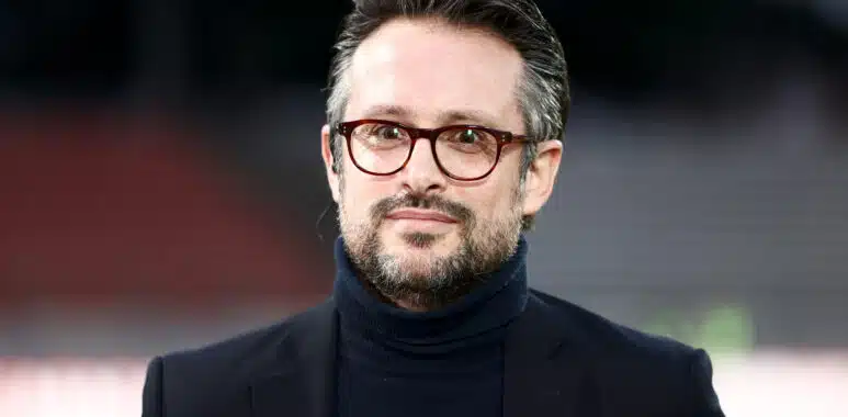 Football Club dAnnecy v Valenciennes Football Club - Ligue 2 BKT | AllezPaillade.com Man wearing a dark blazer and turtleneck with red-framed glasses, outdoors, smiling softly (close-up headshot)