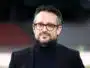 Man wearing a dark blazer and turtleneck with red-framed glasses, outdoors, smiling softly (close-up headshot)