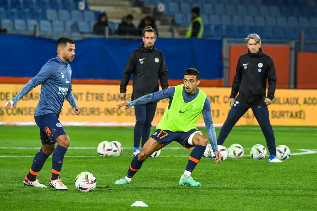 Soccer players train on a grass field, dribbling and passing with multiple balls; foreground player in a neon vest controls the ball.