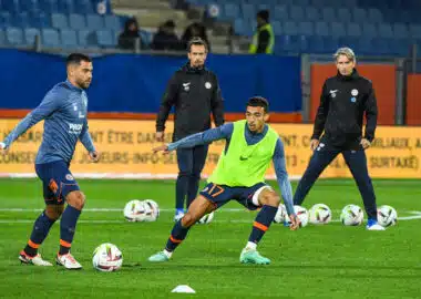 Soccer players train on a grass field, dribbling and passing with multiple balls; foreground player in a neon vest controls the ball.