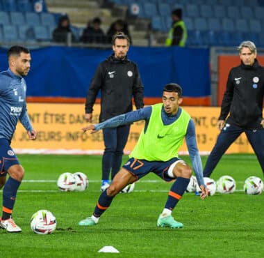 Soccer players train on a grass field, dribbling and passing with multiple balls; foreground player in a neon vest controls the ball.