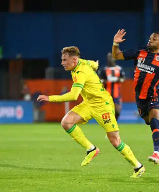 Two soccer players chase the ball on a green field as a referee watches in the background.
