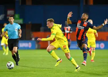 Two soccer players chase the ball on a green field as a referee watches in the background.