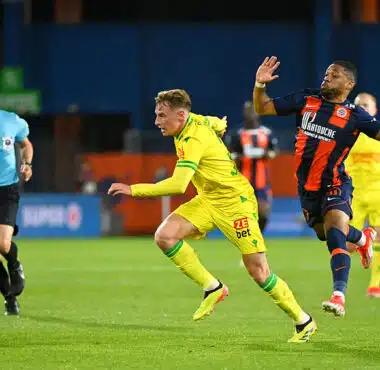 Two soccer players chase the ball on a green field as a referee watches in the background.