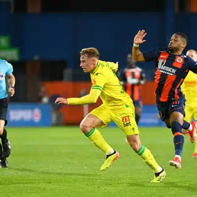 Two soccer players chase the ball on a green field as a referee watches in the background.