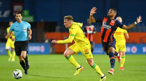 Two soccer players chase the ball on a green field as a referee watches in the background.