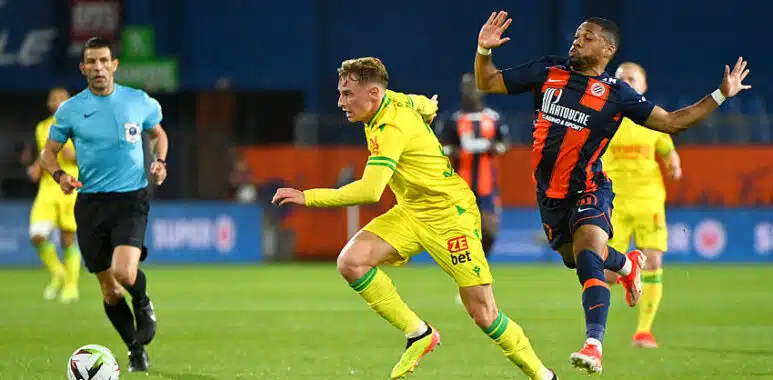 Two soccer players chase the ball on a green field as a referee watches in the background.