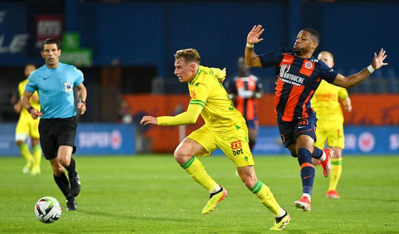 Two soccer players chase the ball on a green field as a referee watches in the background.