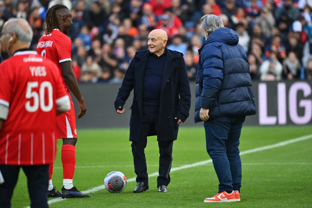 Soccer players in red uniforms stand near a ball on the sideline as two men in dark jackets chat on the field.