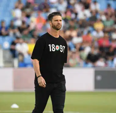 Athlete in a black T-shirt with white numbers and a circular emblem, standing on a field with blurred stadium crowd behind him