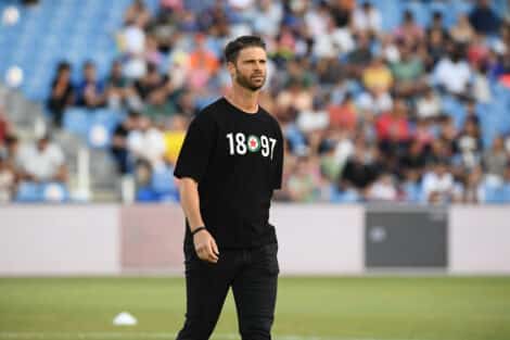 Athlete in a black T-shirt with white numbers and a circular emblem, standing on a field with blurred stadium crowd behind him