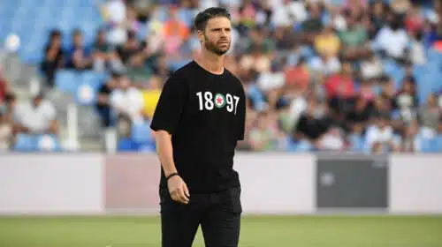 Athlete in a black T-shirt with white numbers and a circular emblem, standing on a field with blurred stadium crowd behind him