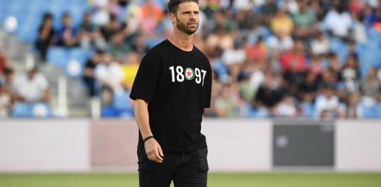 Athlete in a black T-shirt with white numbers and a circular emblem, standing on a field with blurred stadium crowd behind him