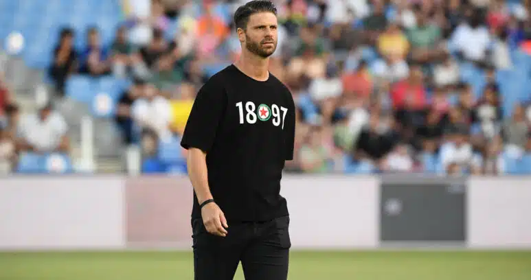 Athlete in a black T-shirt with white numbers and a circular emblem, standing on a field with blurred stadium crowd behind him