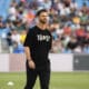 Athlete in a black T-shirt with white numbers and a circular emblem, standing on a field with blurred stadium crowd behind him