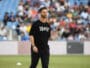 Athlete in a black T-shirt with white numbers and a circular emblem, standing on a field with blurred stadium crowd behind him