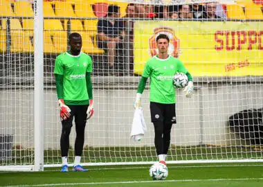 Two soccer goalkeepers in bright green jerseys stand near the goal during training, one holding a ball and a towel.