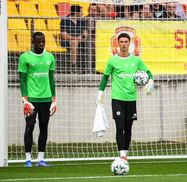 Two soccer goalkeepers in bright green jerseys stand near the goal during training, one holding a ball and a towel.