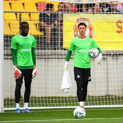 Two soccer goalkeepers in bright green jerseys stand near the goal during training, one holding a ball and a towel.