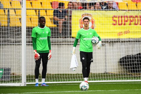 Two soccer goalkeepers in bright green jerseys stand near the goal during training, one holding a ball and a towel.