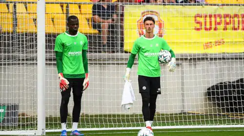 Two soccer goalkeepers in bright green jerseys stand near the goal during training, one holding a ball and a towel.