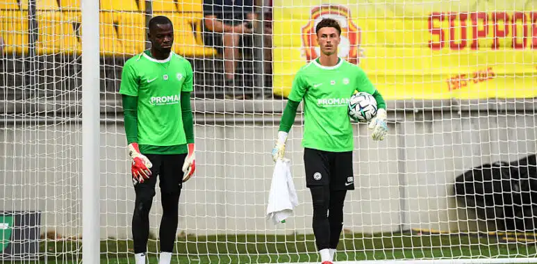 Simon Ngapandouetnbu Dzodic | AllezPaillade.com Two soccer goalkeepers in bright green jerseys stand near the goal during training, one holding a ball and a towel.