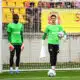 Two soccer goalkeepers in bright green jerseys stand near the goal during training, one holding a ball and a towel.