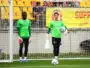 Two soccer goalkeepers in bright green jerseys stand near the goal during training, one holding a ball and a towel.