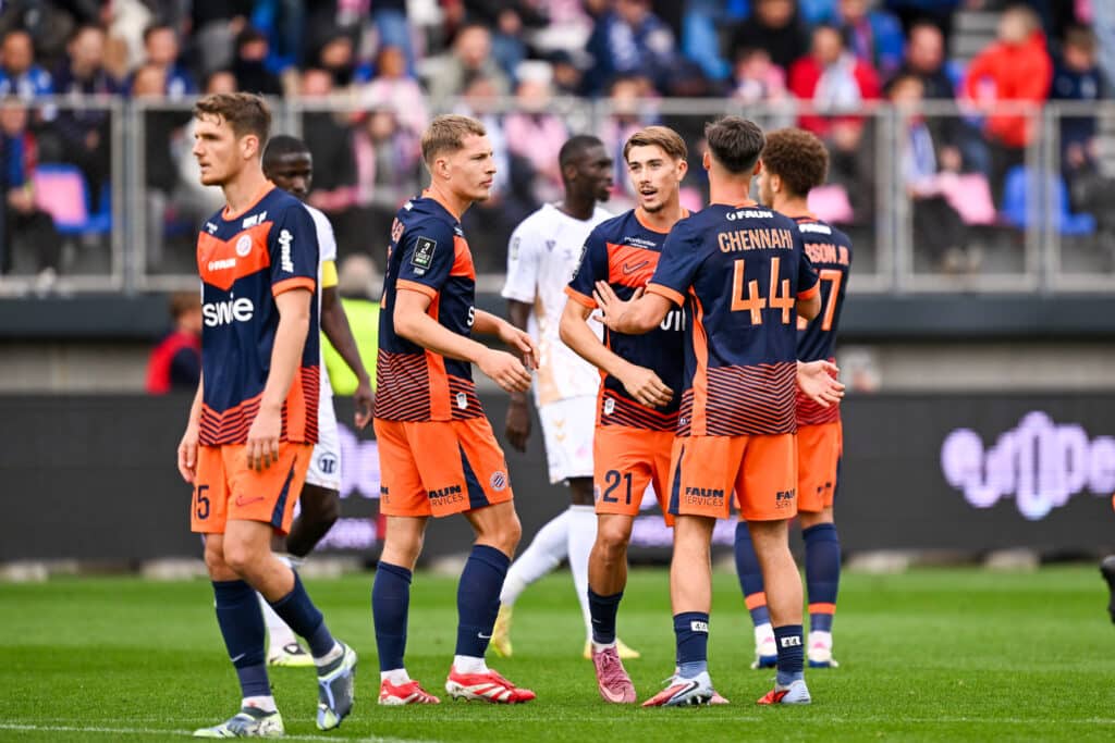 Omeragic Mincarelli Chennahi USL Dunkerque v Montpellier Herault SC - Ligue 2 BKT 20252026 | AllezPaillade.com Soccer players in orange and navy kits gather on a green field, crowd blurred in the background as they celebrate or converse.
