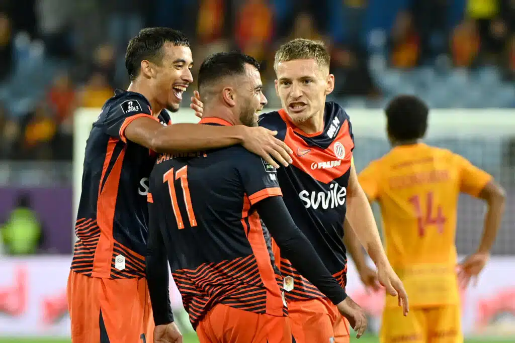 Soccer players in navy and orange jerseys celebrate with a group hug on the field.