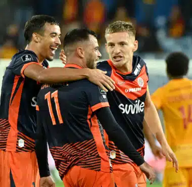 Soccer players in navy and orange jerseys celebrate with a group hug on the field.