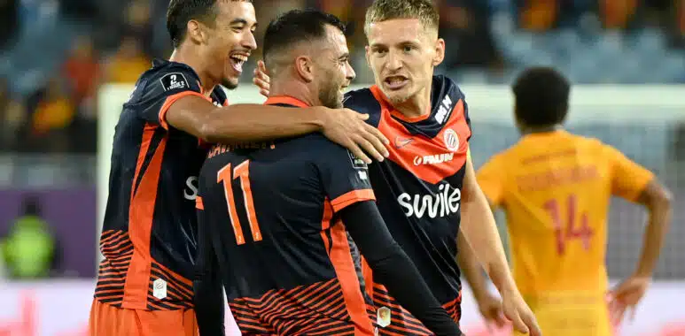 Soccer players in navy and orange jerseys celebrate with a group hug on the field.