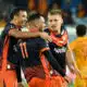 Soccer players in navy and orange jerseys celebrate with a group hug on the field.