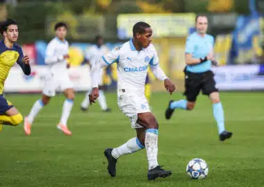 Soccer player in a white and blue kit dribbles a ball on green field during a match, with teammates and a referee in the background.
