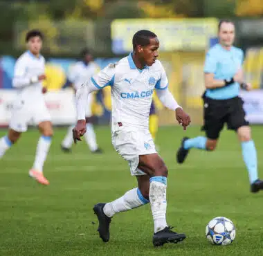 Soccer player in a white and blue kit dribbles a ball on green field during a match, with teammates and a referee in the background.