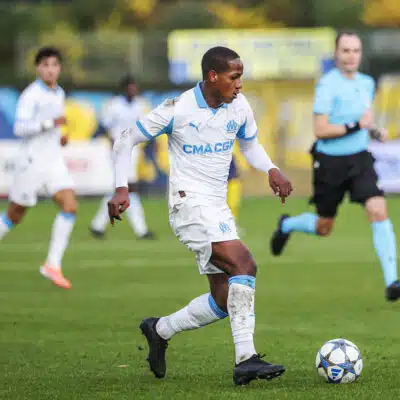 Soccer player in a white and blue kit dribbles a ball on green field during a match, with teammates and a referee in the background.
