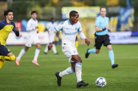 Soccer player in a white and blue kit dribbles a ball on green field during a match, with teammates and a referee in the background.