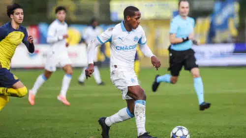 Soccer player in a white and blue kit dribbles a ball on green field during a match, with teammates and a referee in the background.
