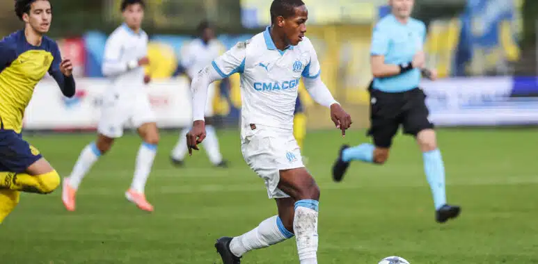 Soccer player in a white and blue kit dribbles a ball on green field during a match, with teammates and a referee in the background.