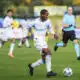 Soccer player in a white and blue kit dribbles a ball on green field during a match, with teammates and a referee in the background.