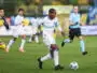 Soccer player in a white and blue kit dribbles a ball on green field during a match, with teammates and a referee in the background.