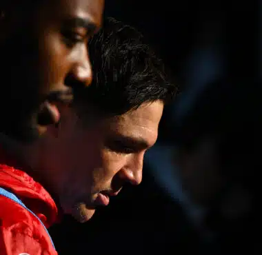 Two athletes standing close with heads bowed; one in a red jacket and the other in a green bib, against a dark background.