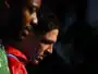 Two athletes standing close with heads bowed; one in a red jacket and the other in a green bib, against a dark background.